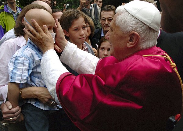 Papst Benedikt XVI. segnet am 19. August 2005, während des XX. Weltjugendtags in Köln, vor der Kirche Sankt Pantaleon die Gläubigen. Er berührt mit den Händen das Gesicht eines krebskranken Kindes und segnet den Jungen.