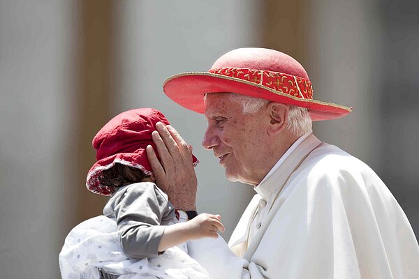 Papst Benedikt XVI. hält behütet den Kopf eines Kleinkindes. Beide tragen einen roten Hut.