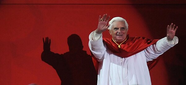 Papst Benedikt XVI winkt sehr freudig. Er steht vor einer komplett roten Wand.